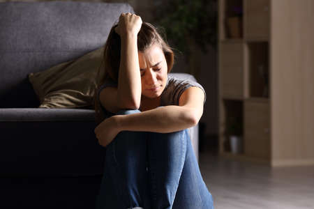Front view of a single sad teen sitting on the floor in the living room at home with a dark backgroundの写真素材
