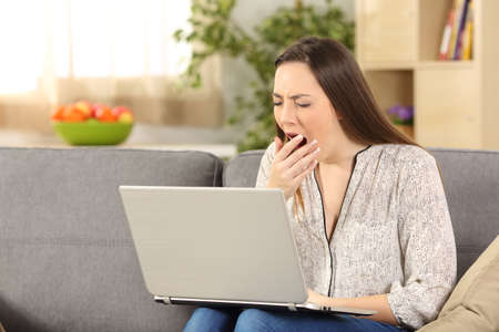 Bored woman on line yawning sitting on a sofa in the living room at homeの写真素材