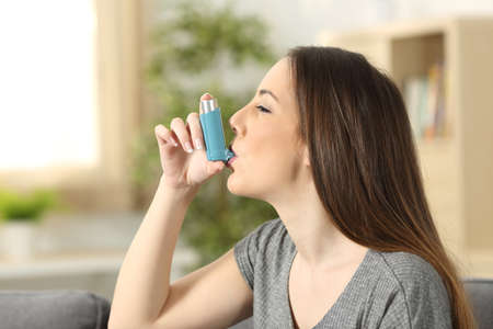 Side view of an asthmatic woman using an inhaler sitting on a couch in the living room at homeの写真素材