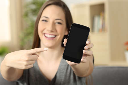 Front view of a girl pointing and showing a blank phone screen looking to you sitting on a sofa in...