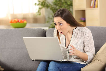 Amazed woman reading on line news in a laptop sitting on a couch in the living room in a house interiorの写真素材