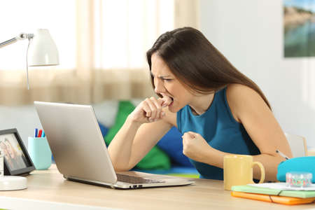 Angry student with a laptop failure sitting in a desk in her room in a house interior with a window in the background