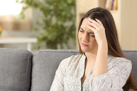 Lady suffering head ache sitting on a sofa in the living room at homeの写真素材