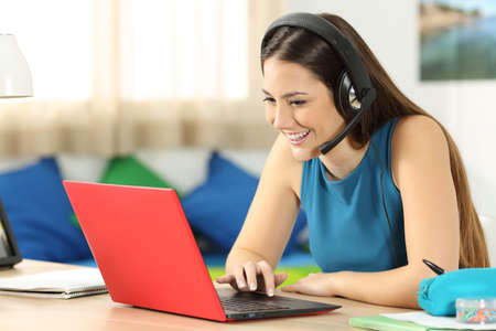 Casual student talking with headsets and a red laptop in a video conference on line sitting in a desk in her bedroomの写真素材