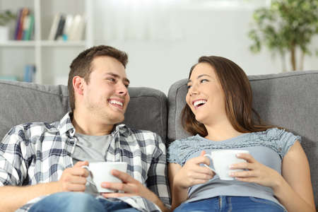 Relaxed couple talking and holding coffee cups sitting on a sofa in the living room at homeの写真素材
