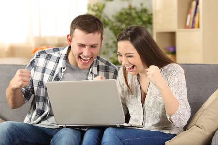 Excited couple reading news on line sitting on a couch in the living room at homeの写真素材
