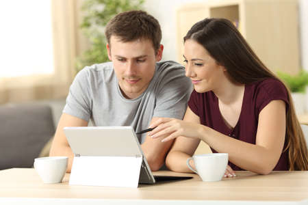 Couple searching on line content with a tablet pc sitting in a table in the living room at home with a window in the background