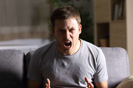 Casual furious man shouting and looking at you sitting on a sofa at home with a dark light in the backgroundの写真素材