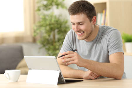 Casual man searching on line using a tablet with a pen sitting in a desk at home with a window in the backgroundの写真素材