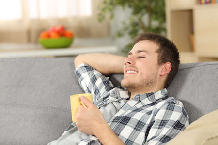 Single relaxed man holding a coffee cup sitting on a sofa in the living room at homeの写真素材