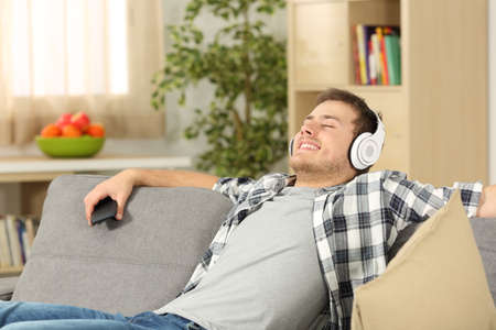 Single casual man with eyes closed listening to music on line from a smart phone sitting on a sofa in the living room in a house interior