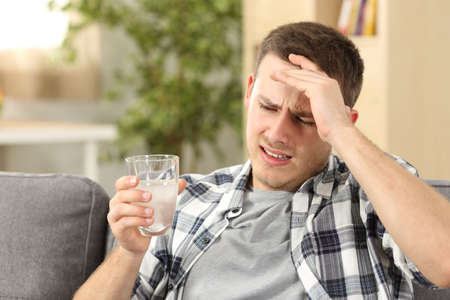 Man suffering head ache with a hand on forehead and holding a glass with a painkiller sitting on a couch at homeの写真素材