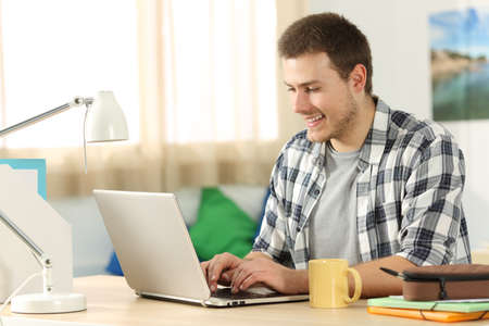 Happy student writing in a laptop sitting on a desk in his roomの写真素材