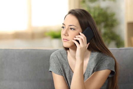 Serious woman attending a phone call sitting on a sofa in the living room at homeの写真素材