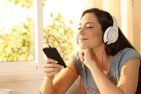Relaxed woman listening music with headphones and a smart phone at home with a window in the backgroundの写真素材