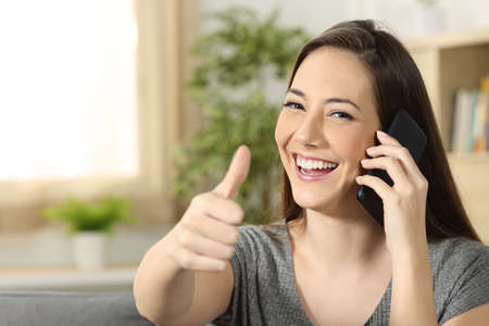 Woman talking on a phone call looking at you sitting on a sofa in the living room at homeの写真素材
