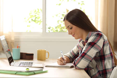 Side view of a single student studying and taking notes in a table at home with a window in the backgroundの写真素材