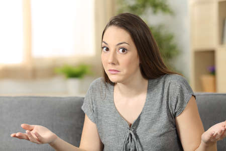 Portrait of a doubtful woman looking at camera sitting on a sofa in the living room of an apartment