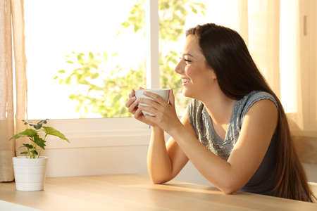 Happy woman relaxing with a coffee cup and looking through a window sitting in a table at homeの写真素材