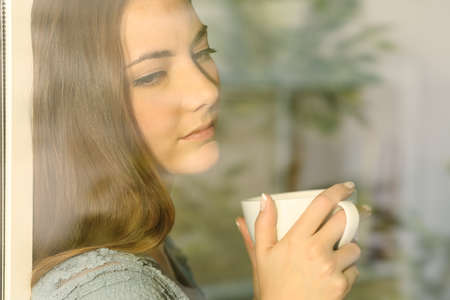 Pensive lady looking through a window holding a cup of coffeeの写真素材