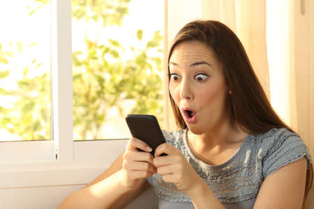 Single amazed girl watching content on a smart phone at homeの写真素材