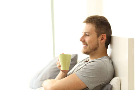 Side view of a happy man holding coffee cup having breakfast and thinking on the bed of an apartmentの写真素材