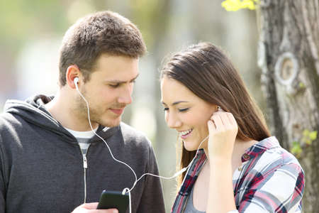 Couple flirting and sharing music on line with earphones and a smart phone outdoors in a parkの写真素材