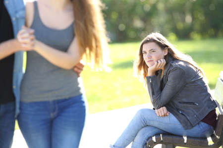 Single envious girl sitting on a bench and seeing an affectionate couple who is walking in a park