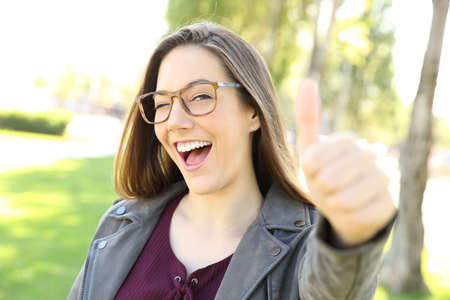 Portrait of a funny girl wearing eyeglasses gesturing thumbs up and looking at you outdoors in a parkの写真素材