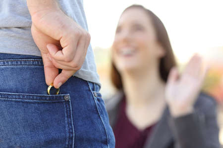 Cheater hiding wedding ring in a pocket in foreground while is dating with his lover outdoors