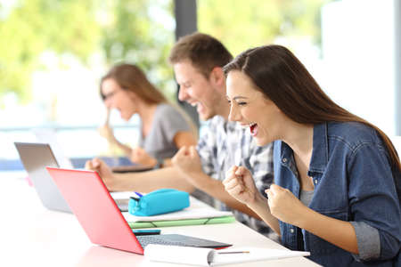 Three excited classmates receiving good news on line in their laptops in a classroomの写真素材