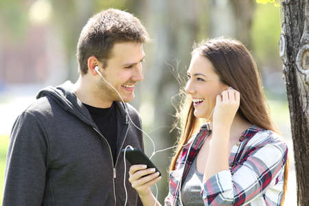Happy couple of teens sharing on line music in a smart phone outdoors in a parkの写真素材