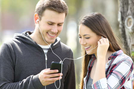 Happy couple of teenagers sharing on line music and watching media content in a smart phone outdoors in a parkの写真素材