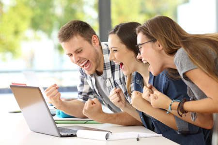 Side view of three excited students reading good news together on line in a laptop sitting in a desk in a classroom