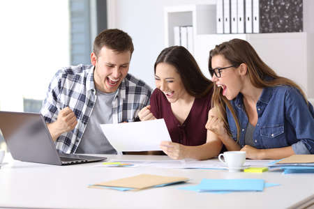 Three excited workers reading a good results report sitting in a desktop at officeの写真素材