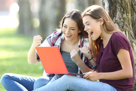 Excited teens receiving good news on line sitting on the grass in a parkの写真素材