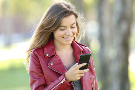 Fashion teen wearing red jacket chatting on line with a mobile phone walking in a parkの写真素材