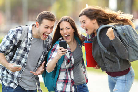 Front view of three excited students receiving good news on line in a mobile phone in an university campus or street