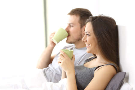 Side view of a relaxed couple having breakfast holding cups on the bed of an hotel room or homeの写真素材