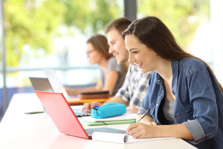 Side view of a student e-learning with a laptop and taking notes on a table in a classroom with a green backgroundの写真素材