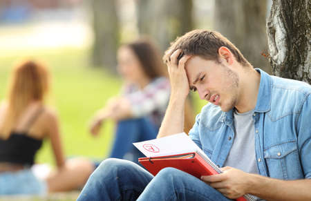 Single sad student checking a failed exam sitting on the grass in a park with unfocused people in the background