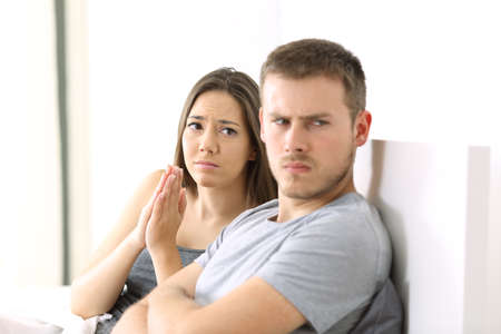 Wife begging to her angry husband sitting on the bed in a house interiorの写真素材