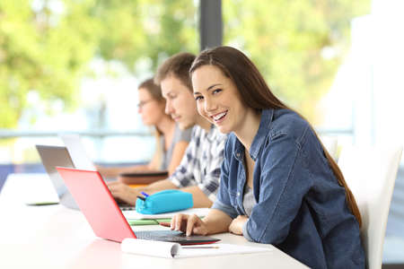 Student girl with a laptop posing and looking at you in a classroom with classmates in the backgroundの写真素材