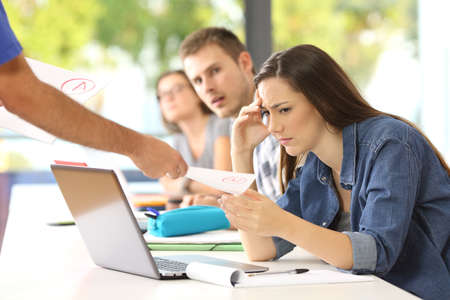 Sad student receiving a failed exam in a classroom with classmates watching in the backgroundの写真素材