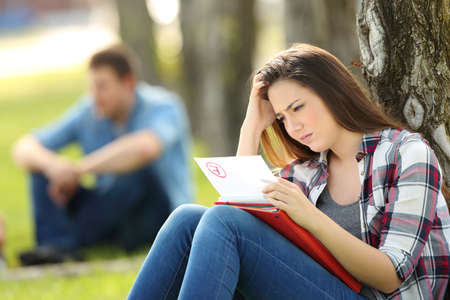 Single sad student looking at failed exam sitting on the grass in a park with unfocused people in the backgroundの写真素材