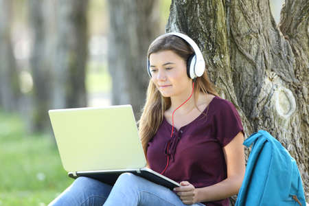 Single teen girl studying watching video tutorials on line in a laptop sitting on the grass leaning in a tree in a parkの写真素材