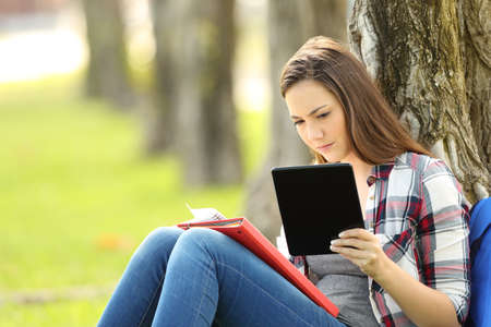 Single student studying comparing notes on line with a tablet sitting in a park leaning on a treeの写真素材