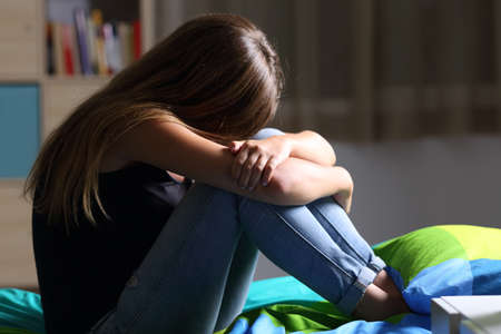 Portrait of a single sad teen sitting on the bed in her bedroom with a dark light in the background