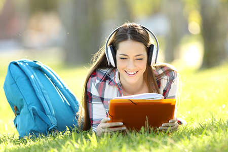 Front view portrait of a student reading notes and listening an audio tutorial on line lying on the grass in a parkの写真素材