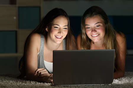 Two teens watching on line content in a laptop lying on the floor in a room at home with a dark light in the backgroundの写真素材